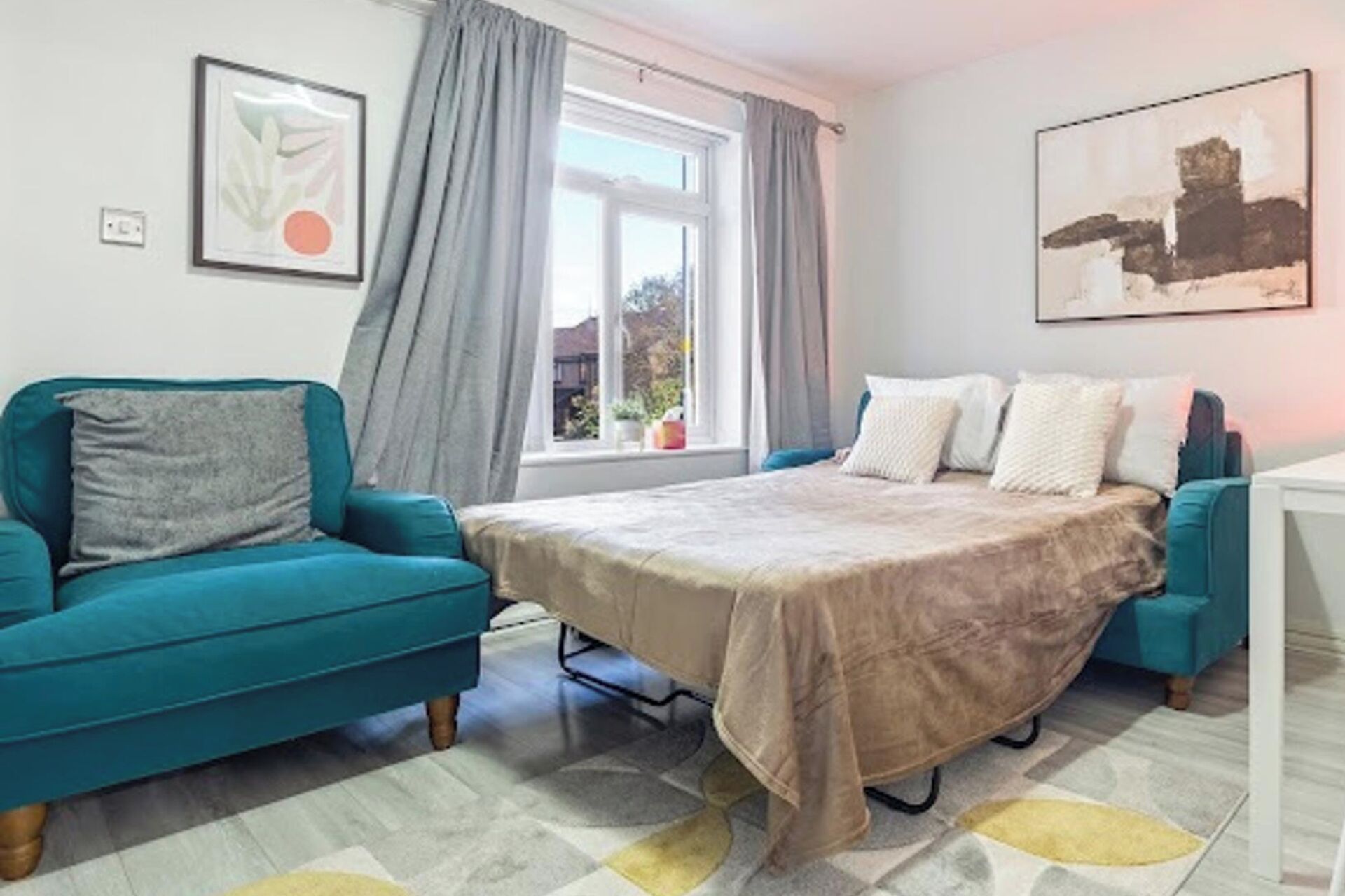 A bedroom with a bed, a teal sofa, a window with curtains, and two framed pictures on the wall.