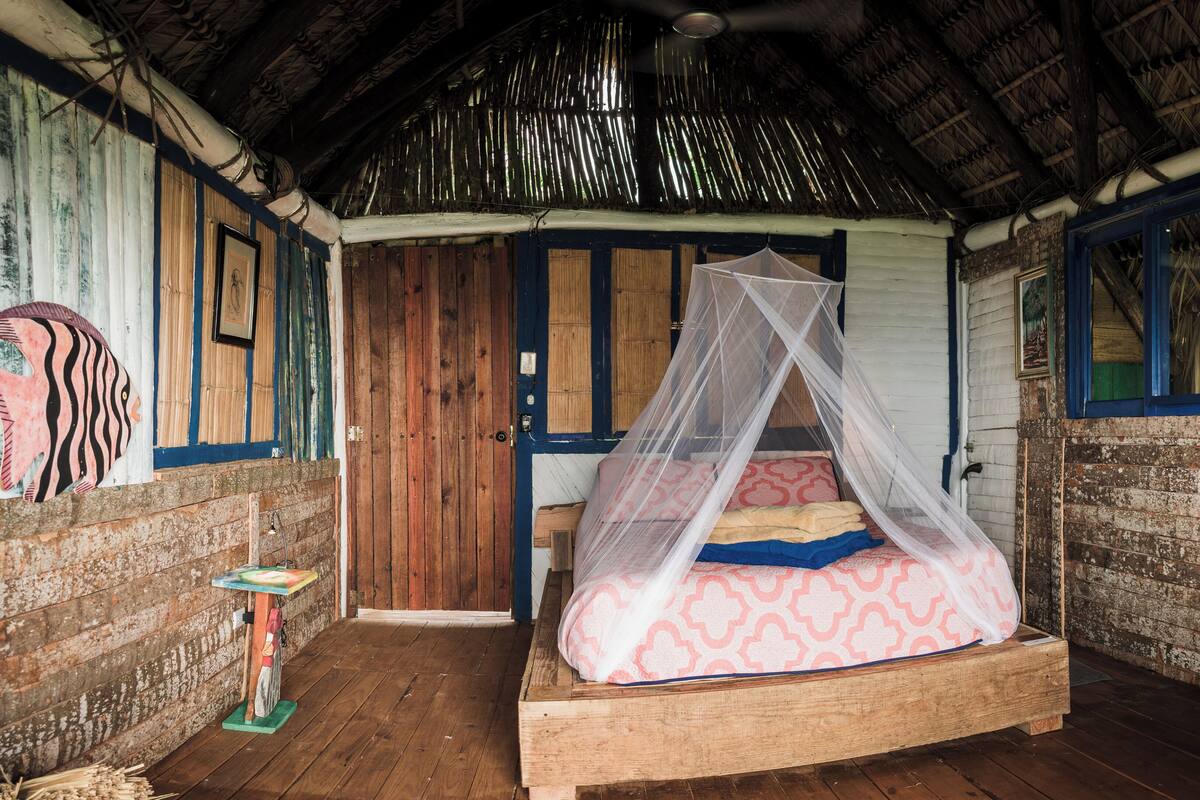 A rustic room with a wooden bed, a mosquito net, and a thatched roof.