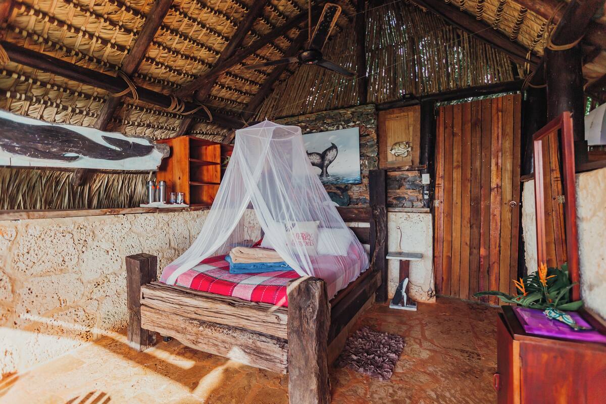 A rustic wooden bed with a mosquito net, a stone wall, a wooden shelf with bottles, and a wooden door.