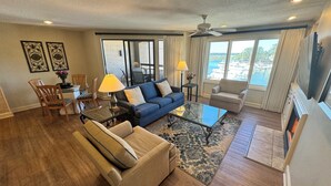 A spacious living room with a blue sofa, beige armchair, and a glass coffee table. There is a dining area with a table and chairs, and a large window offering a view of the ocean. - Gorgeous 2 bedroom villa overlooking Shelter Cove Harbour with pool and spa (Hilton Head Island)