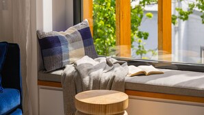 En mysig läshörna med en bekväm sittplats, ett litet träbord och ett fönster med utsikt över grönskan.