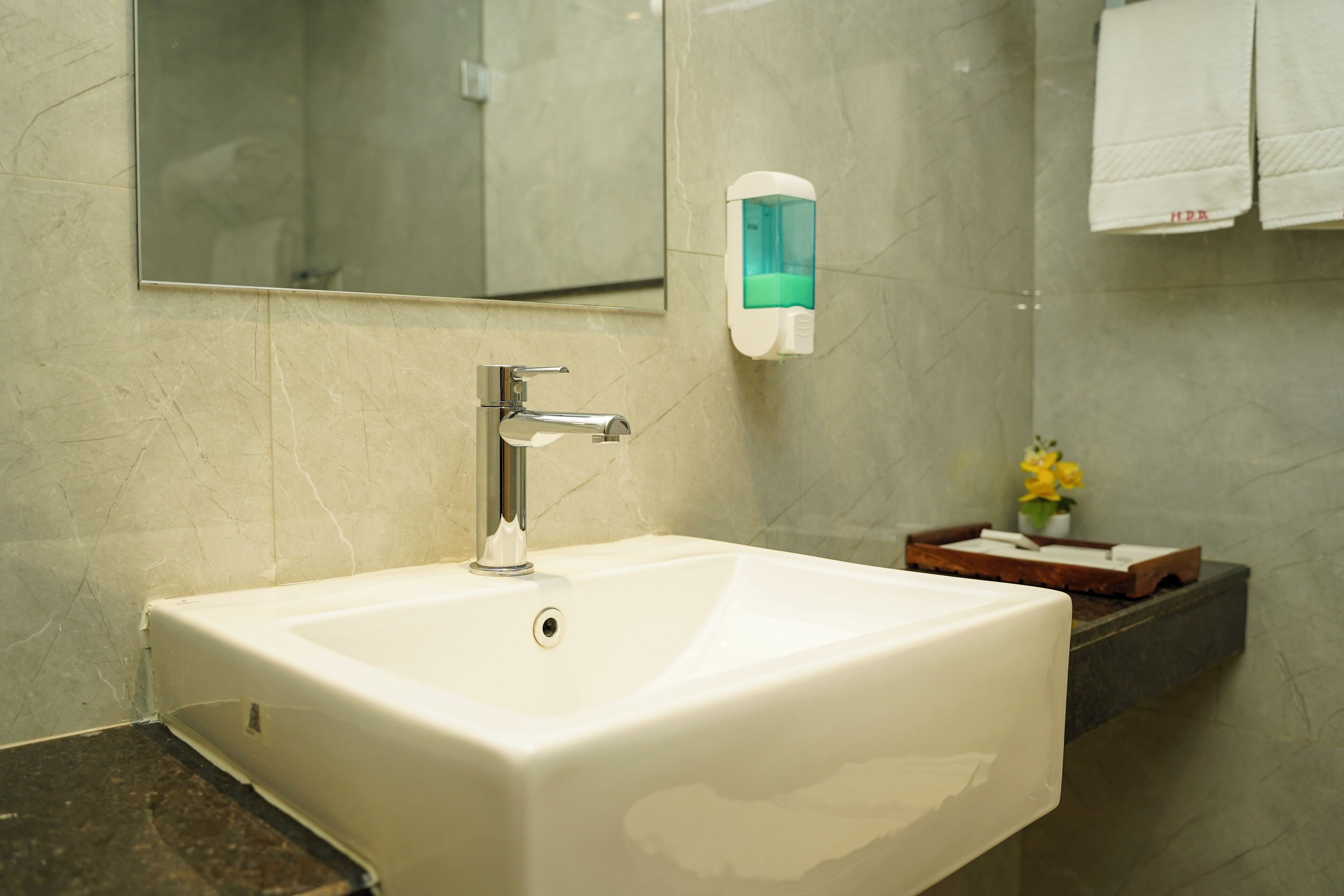 A bathroom with a white sink, a mirror, a soap dispenser, and a small shelf with a vase of yellow flowers.