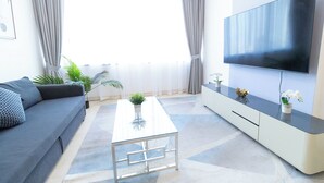 Een moderne woonkamer met een grijze bank, een glazen salontafel, een tv op een kast en een groot raam met gordijnen.