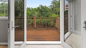 An open glass door leading to a wooden deck with a metal railing, surrounded by greenery.