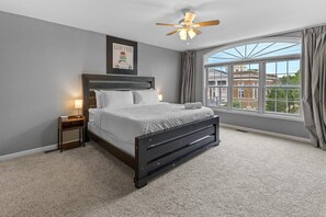 A bedroom with a bed, a nightstand, a ceiling fan, a window with curtains, and a view of buildings outside. - DT Indy near Convention Center (Indianapolis)