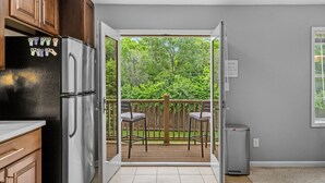 A kitchen with a refrigerator, a dining area with chairs, a wooden deck, and a view of greenery.