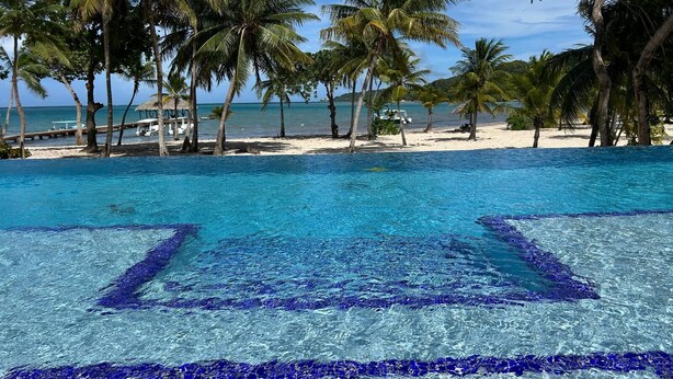 Une piscine d’un bleu éclatant, bordée d’une terrasse carrelée, entourée de palmiers et donnant sur une plage.
