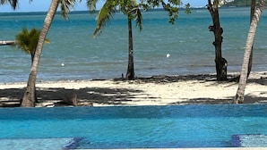Une piscine à l’eau d’un bleu cristallin, entourée de dalles de pierre, de palmiers et d’une plage de sable fin, avec l’océan en toile de fond.