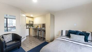 A compact hotel room with a kitchenette, a bed with striped bedding, a leather chair, and a view of the city through a window.