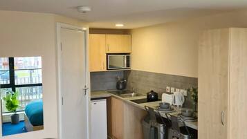 A compact kitchen with a microwave, sink, and a small refrigerator. There are two bar stools and a black chair. The kitchen has wooden cabinets and a window with a view of a balcony.