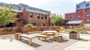 Un espace extérieur aménagé en terrasse, avec des bancs et des tables en bois, entouré de bâtiments en briques et de verdure.