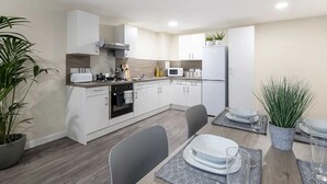 Une cuisine moderne avec des armoires blanches, un four encastré, un four à micro-ondes et un réfrigérateur. On y trouve une table à manger avec des chaises et une plante en pot.