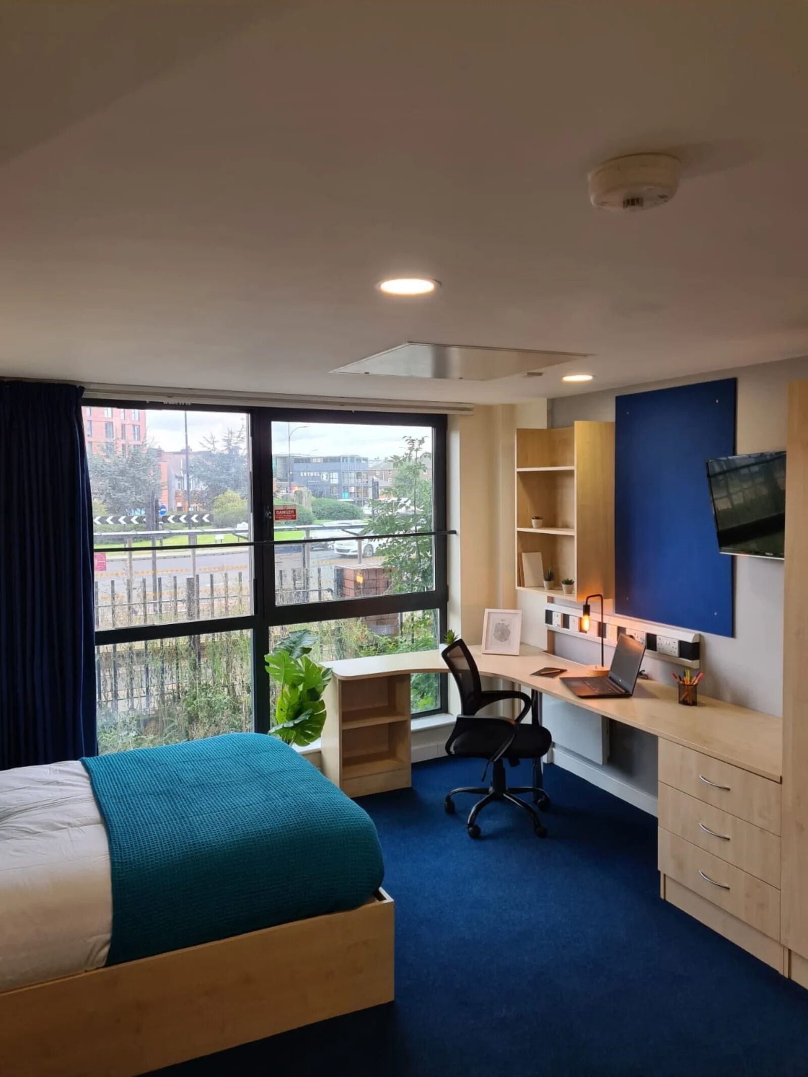 A modern bedroom with a bed, desk, chair, and a large window offering a city view.