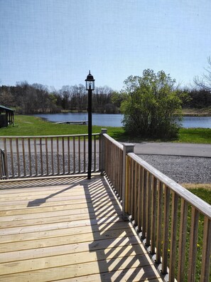 Une terrasse en bois avec une rambarde en métal donnant sur un lac, un lampadaire et une zone herbeuse avec des arbres.
