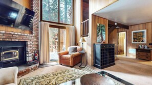 A cozy living room with a fireplace, leather sofa, and a large window with a view of trees. - Large Townhouse, Fireplace, Walk to the Winter Shuttle, Ice Rink & Bowling (Mammoth Lakes)