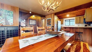 Une table de salle à manger en bois, agrémentée d’un set de table, un lustre et un coin cuisine avec des meubles en bois et une fenêtre.