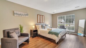A bedroom with a bed, a sofa, a nightstand, a lamp, a window with blinds, and a plant.