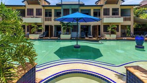 Zona de piscina con una sombrilla azul, sillones de descanso y un edificio de dos plantas con balcones.