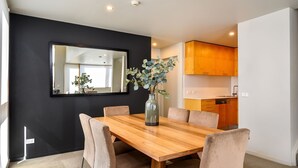 A modern dining area with a wooden table, six chairs, and a vase with green foliage. A large mirror is mounted on the wall, and a kitchen with wooden cabinets is visible in the background. - Snow Stream 5 a 2 bedroom & loft apartment close to Friday Flat. (Thredbo)