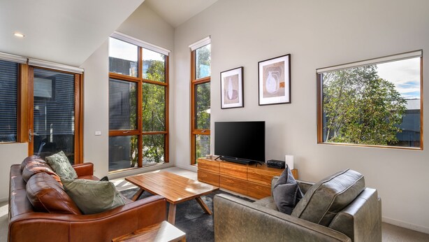 A spacious living room with a large TV, wooden furniture, and large windows offering views of trees and buildings. - Snow Stream 5 a 2 bedroom & loft apartment close to Friday Flat. (Thredbo)