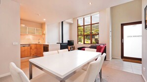 A modern kitchen with wooden cabinets and a built-in oven, a dining area with a white table and chairs, and a large window offering a view of greenery.