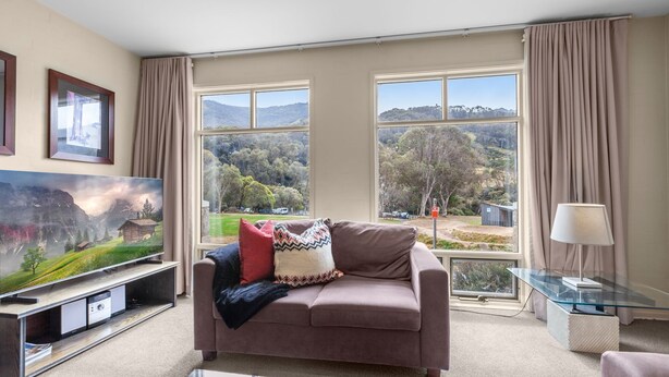 A living room with a large flat-screen TV, a brown sofa, and a glass coffee table. - Village Green Townhouse 2 bedrooms and loft with spectacular views, garage parking and a wood fire. (Kosciuszko National Park)