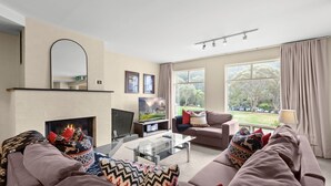 A living room with a fireplace, a television, and a sofa with decorative pillows. - Village Green Townhouse 2 bedrooms and loft with spectacular views, garage parking and a wood fire. (Kosciuszko National Park)