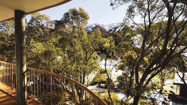 Una terraza de madera con baranda de metal que da a un paisaje nevado con árboles y una montaña al fondo.