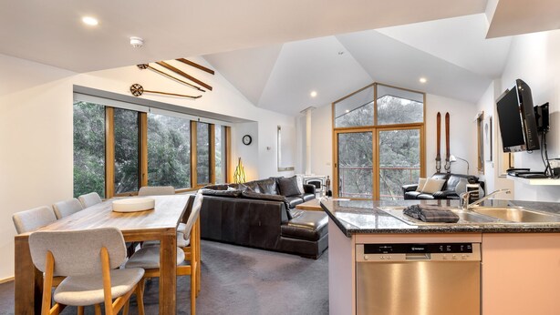 Una cocina moderna con lavavajillas, un comedor con mesa y sillas, un sofá y ventanas amplias con vistas a árboles.