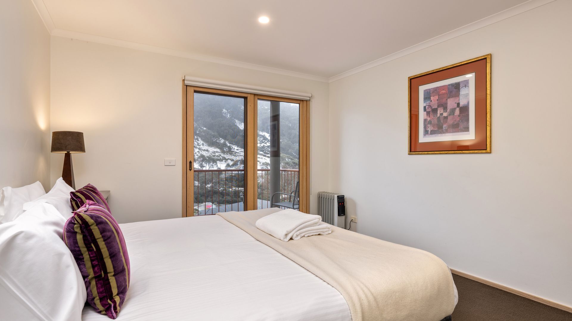 A hotel room with a bed, a window with a view of snow-capped mountains, a radiator, and a framed picture on the wall.