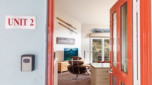 Una habitación con un cartel que dice “UNIT 2”, un escritorio con una silla, un ventanal con vista a un paisaje montañoso y un portaequipajes para esquíes.
