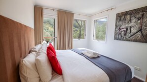 Un dormitorio con una cama grande, cortinas color beige y vistas a árboles a través de las ventanas.