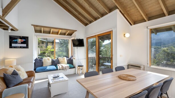 Une pièce chaleureuse avec un plafond en bois, un canapé agrémenté de coussins, un téléviseur fixé au mur et une grande fenêtre donnant sur un paysage verdoyant.
