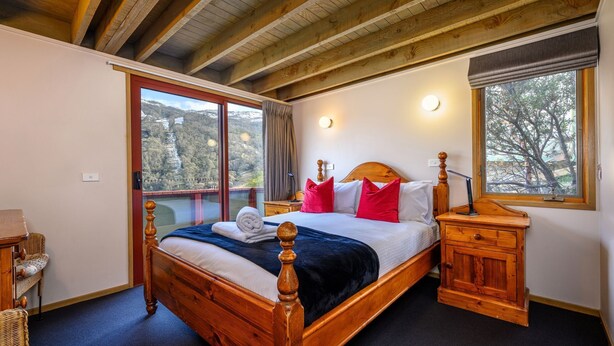 A bedroom with a wooden bed, a nightstand, a wicker chair, a large window with a view of a bridge and trees, and a ceiling with exposed beams. - Banjo 5 - Two Bedroom & Loft Townhouse with mountain views (Thredbo)