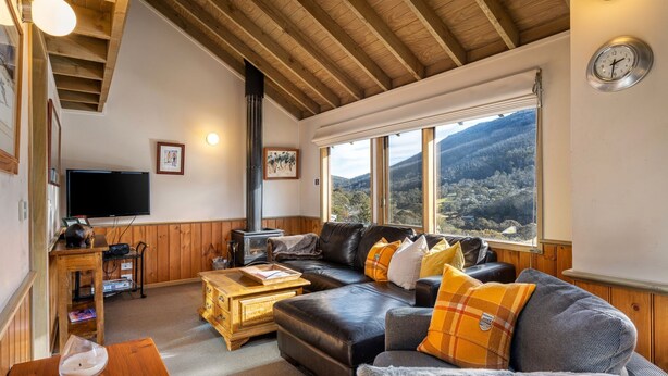 Een gezellige woonkamer met een houten plafond, een open haard, een groot raam met uitzicht op de bergen, een leren bank en een televisie.