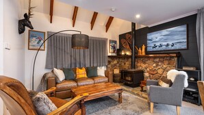 A cozy living room with a stone fireplace, leather sofa, and a large TV mounted on the wall. - A stand alone 2 bedroom chalet with a large veranda to take in the mountain views (Thredbo)