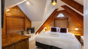 A bedroom with a sloped ceiling, wooden beams, a bed with white bedding, two lamps, and a wooden desk. - A stand alone 2 bedroom chalet with a large veranda to take in the mountain views (Thredbo)