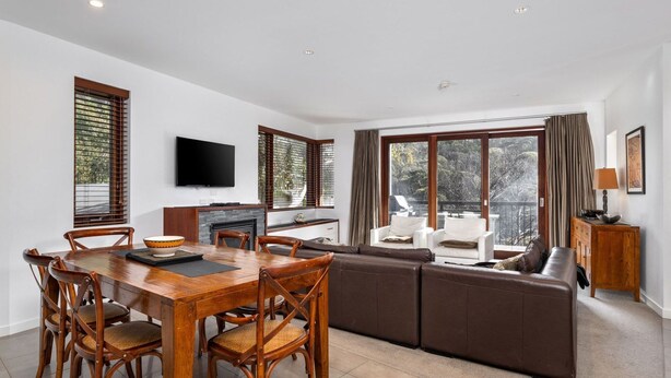 A modern living room with a wooden dining table, comfortable seating, a fireplace, and large windows offering a view of the outdoors.