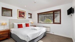 A modern hotel room with a large bed, two bedside tables with lamps, a flat-screen TV, and large windows with blinds.