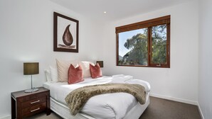 Un dormitorio con cama, mesita de noche, lámpara, un cuadro enmarcado y ventana con vistas a árboles.