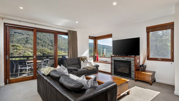 Amplia sala de estar con sofá de cuero, televisor de pantalla plana, chimenea y ventanas amplias que ofrecen una vista panorámica de las montañas.