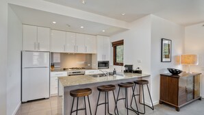 Una cocina moderna con armarios blancos, encimera de granito, horno integrado, microondas y cafetera. Hay cuatro taburetes de madera en la isla.