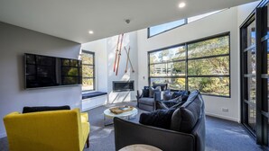 A modern living room with a large TV, comfortable seating, and large windows offering a view of the outdoors.