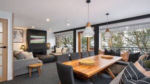 Een moderne woonkamer met een open haard, een eethoek en grote ramen met uitzicht op bomen.