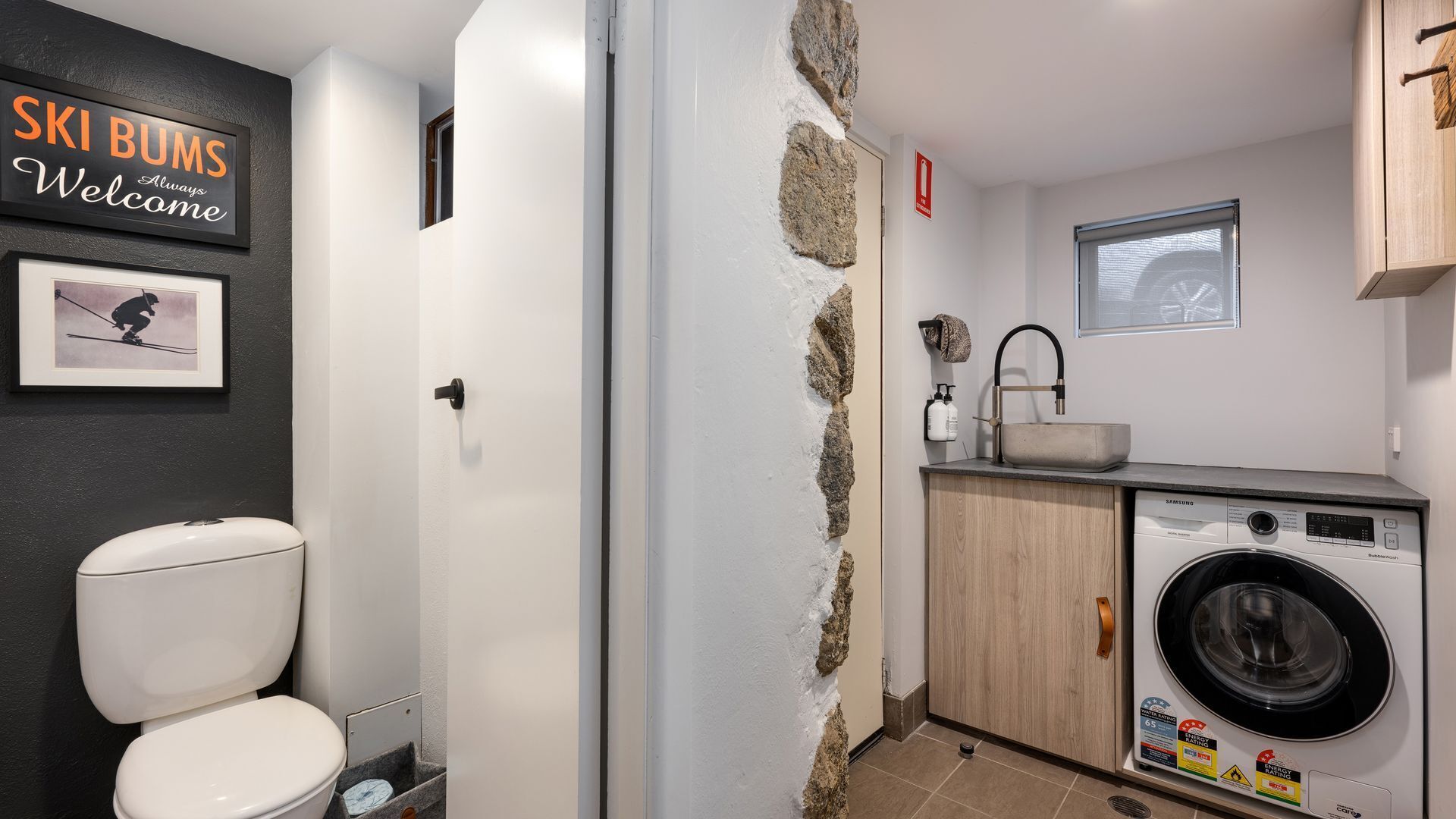 Une petite salle de bain équipée de toilettes, d’un lave-linge et d’un lavabo. Un panneau est accroché au mur avec l’inscription suivante : « Les amateurs de ski sont les bienvenus ».