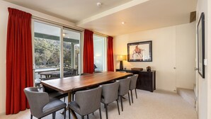 A dining area with a wooden table and six chairs, red curtains, a framed picture on the wall, and a view of a balcony with a table and chairs outside.