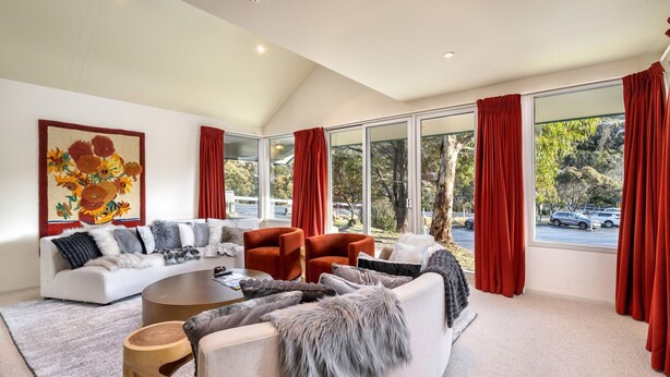 A modern living room with a white sofa, red velvet curtains, and a large window offering a view of a parking lot and trees.