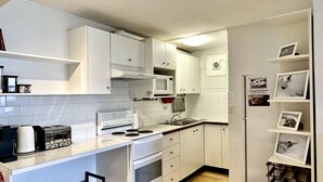 Una cocina funcional con armarios blancos, microondas, estufa y lavamanos. Se observan estantes con fotos enmarcadas y un refrigerador pequeño.