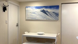Una habitación con una puerta blanca, una cabeza de venado montada en la pared, un cuadro de un paisaje montañoso y una pequeña mesa blanca con un cuenco.