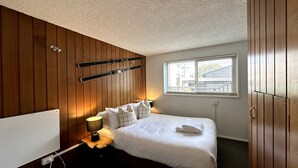 Un dormitorio con pared de paneles de madera, cama con ropa blanca, lámpara de noche y ventana con persianas.